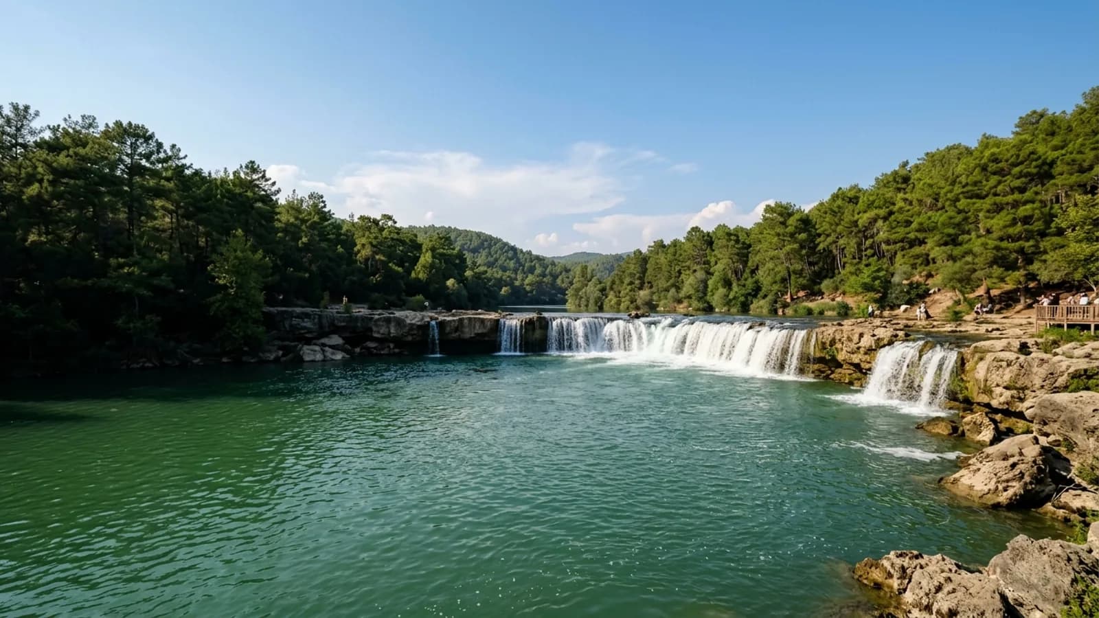 Manavgat, Türkiye — the Manavgat river, a wide low waterfall spilling over a broad rock ledge into green water with pine woods along both banks.