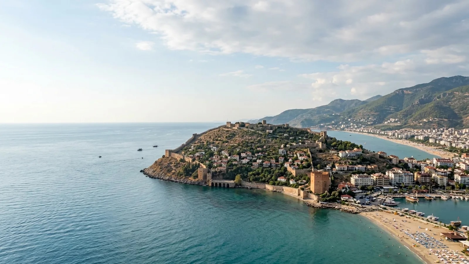 Alanya, Türkiye — the town of Alanya, a rocky peninsula crowned by a medieval Seljuk castle wall with a red brick tower at the harbour and long sandy beaches on both sides.