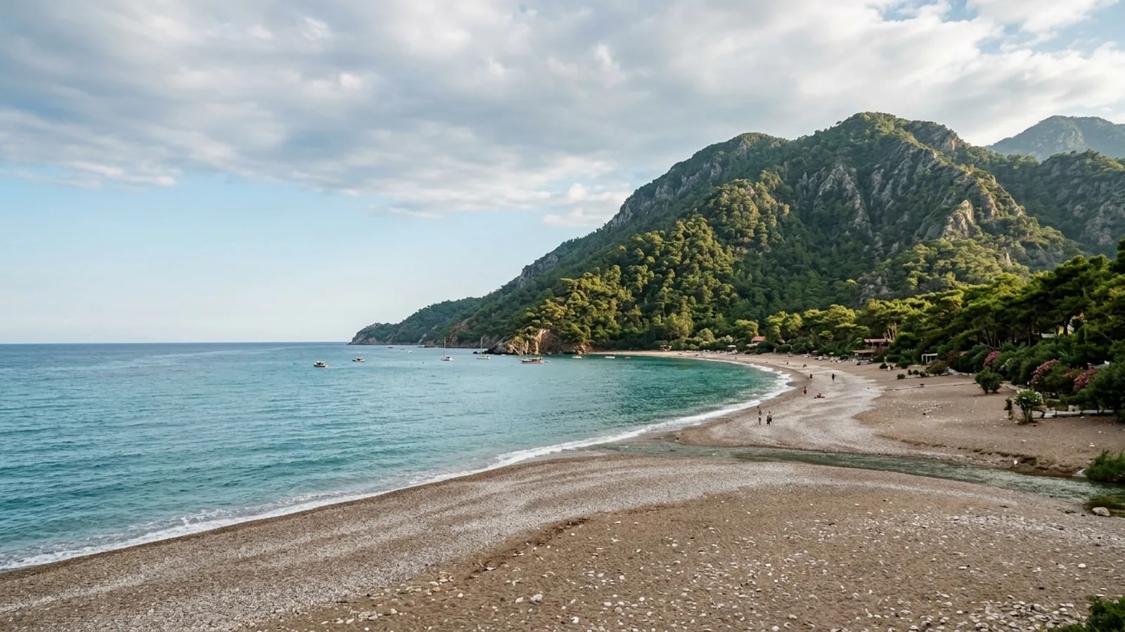 Cirali, Türkiye — the wide pebble beach at Cirali below wooded mountains, a broad open bay with a stream mouth and unspoilt low-rise surroundings.