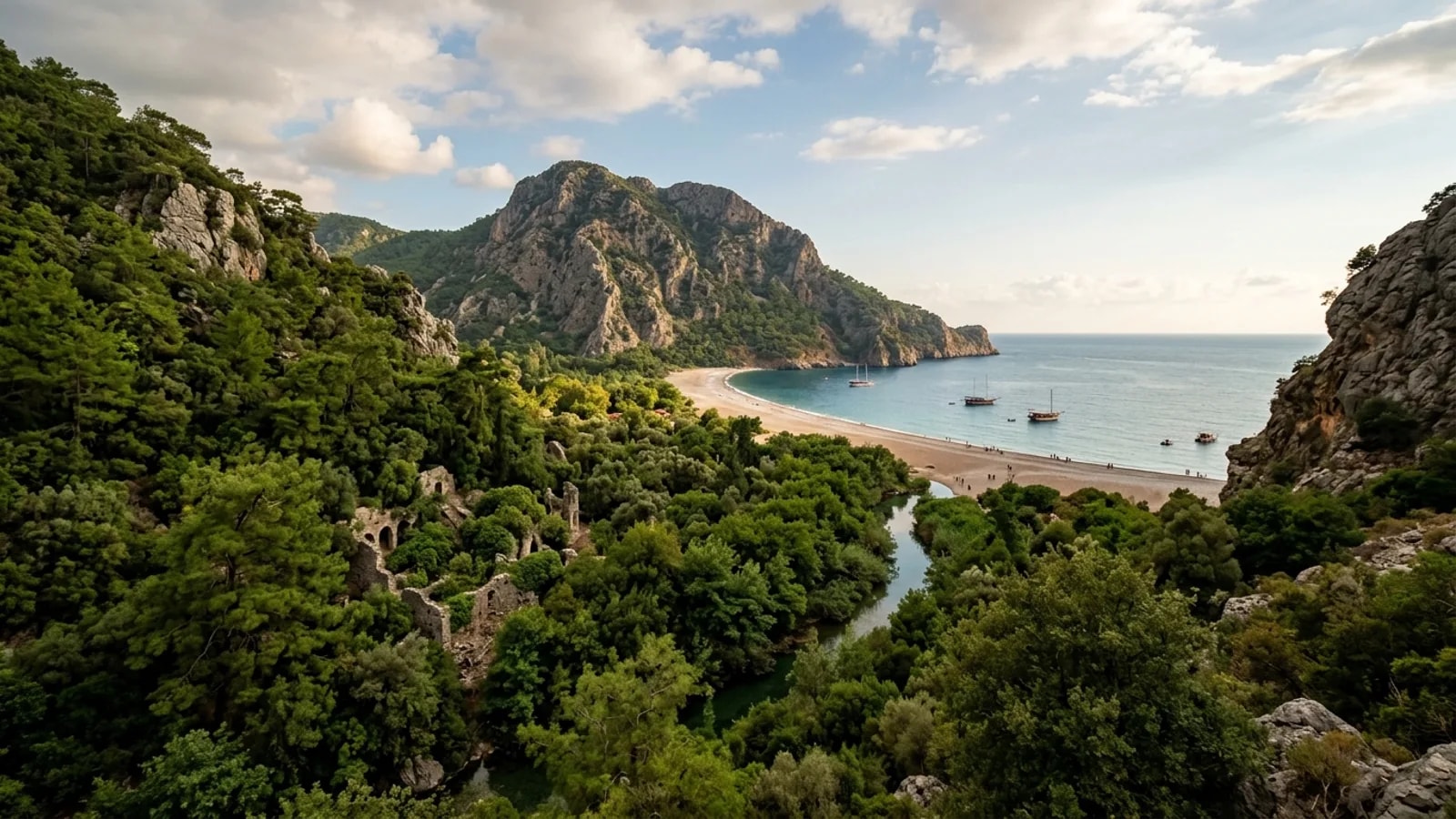 Olympos, Türkiye — the Olympos valley on the Lycian coast, ancient ruins half hidden in dense green woodland along a stream that reaches a long pebble beach between rocky headlands.